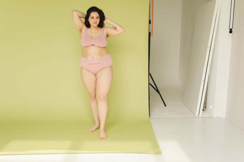 Plus-size woman in lingerie posing confidently in a studio with green background.