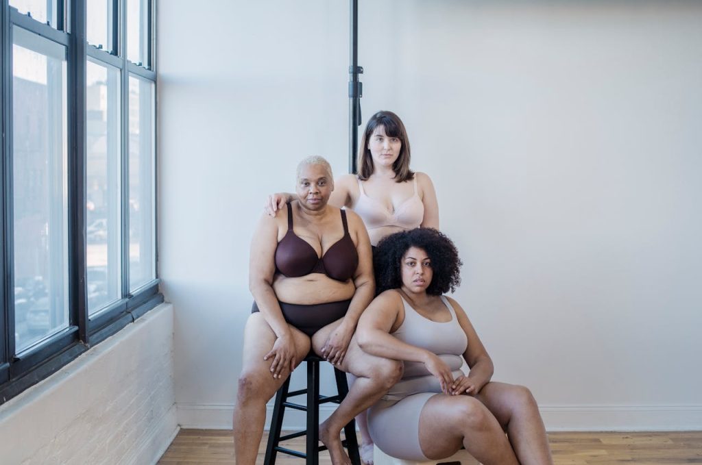 Plump multiracial women in underclothes looking at camera against white wall in building in daylight